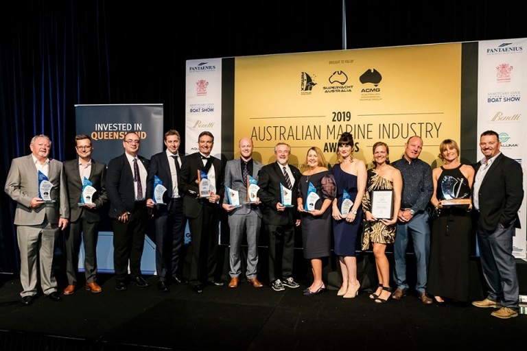 A group of people stand on stage holding trophies and certificates at the 2019 Australian Marine Industry Awards, with banners and a yellow event backdrop behind them.
