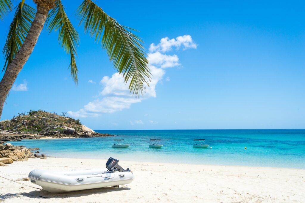A white inflatable boat rests on a sandy tropical beach with clear turquoise water, three small boats anchored offshore, a palm tree in the foreground, and a rocky hill on the left under a sunny blue sky.