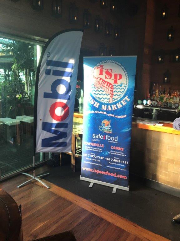 Two vertical banners stand indoors. The left banner displays the Mobil logo. The right banner promotes TSP Fish Market and Safe Food, with images of seafood and contact information. A bar and seating area are in the background.