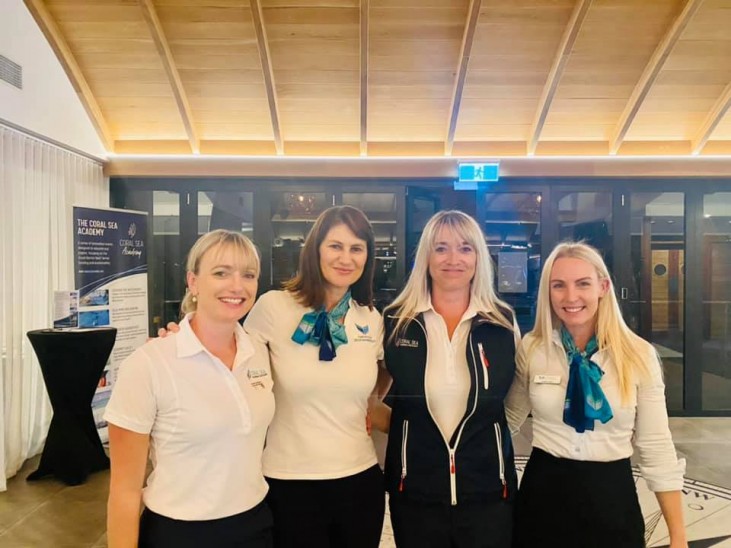 Four women in uniform shirts and neck scarves stand together indoors, smiling at the camera. They are in a well-lit room with a wooden ceiling and glass doors in the background.