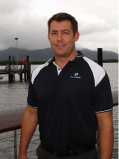 A man in a navy and white collared shirt stands by a waterfront with boats and docks in the background, and misty mountains under a cloudy sky.