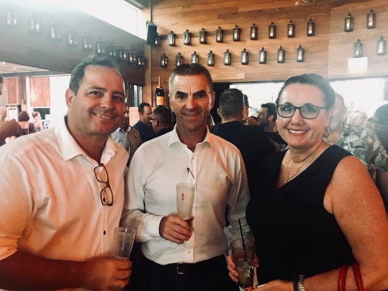Three adults, two men and one woman, stand closely together smiling at an indoor event, each holding a drink. The background shows other people mingling and a wooden wall decorated with lanterns.