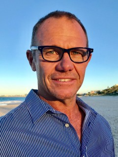 A man wearing glasses and a blue striped shirt smiles whilst standing on a sunny beach with blue sky and distant buildings in the background.
