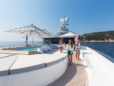 A family of four stands on the deck of a luxury yacht near a swimming pool, surrounded by clear blue water and a rocky shoreline in the background under a sunny sky.