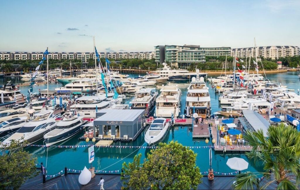 A marina filled with numerous yachts and boats moored side by side, with modern buildings and greenery in the background under a clear sky. Flags and banners decorate the docks, adding a festive atmosphere.