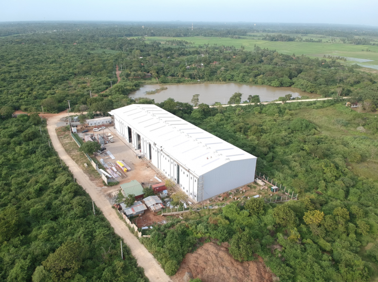 Aerial view of a large white industrial warehouse surrounded by dense green vegetation, with a pond and fields in the background, and a dirt track leading to the facility.