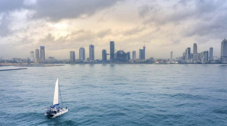 A small sailing boat moves across calm blue water under a cloudy sky, with a modern city skyline full of tall buildings and skyscrapers in the background.