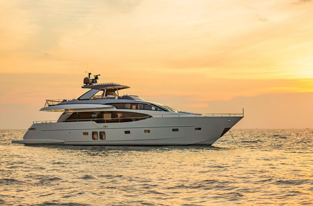 A luxury yacht floats on calm ocean waters during a golden sunset, with the sky glowing in warm orange and yellow hues.