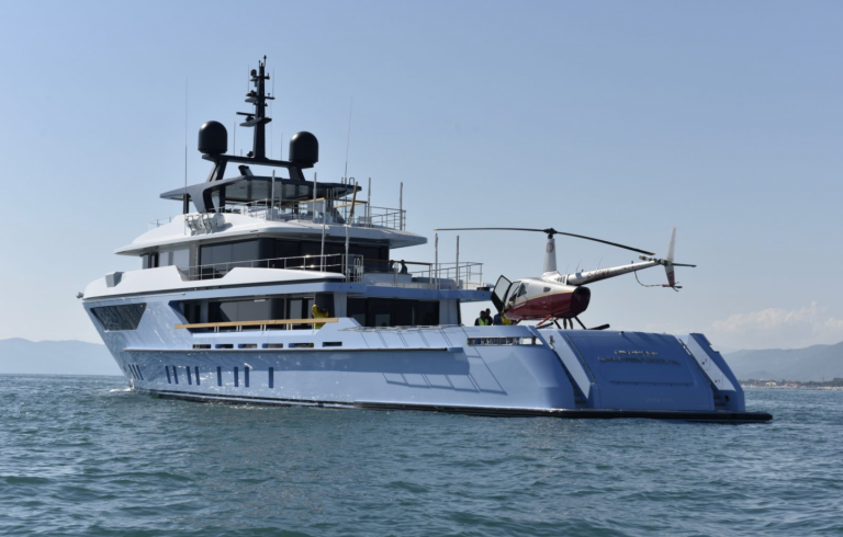 A large modern yacht with a helicopter on its rear deck floats on calm water near a coastline, with mountains visible in the background under a clear sky.