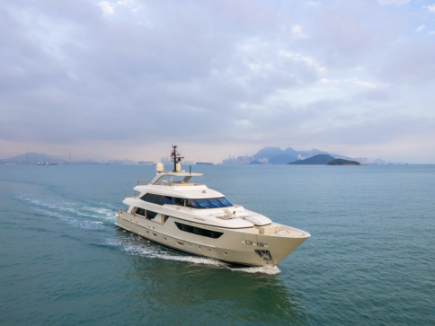 A white luxury yacht cruises through calm blue-green water with an island and distant mountains visible under a partly cloudy sky.
