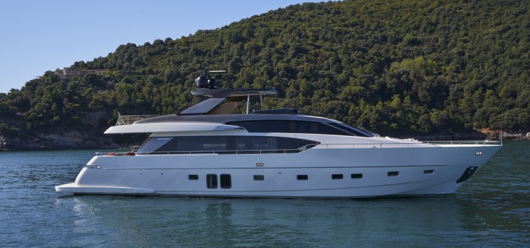 A modern white yacht with sleek design floats on calm blue water, with a lush green forested hill in the background under a clear sky.