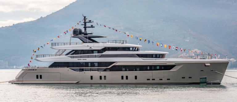 A large, modern yacht decorated with colourful signal flags is moored on calm water, with a mountainous landscape and some scattered buildings in the background.