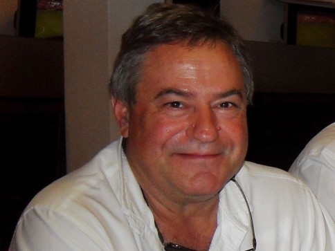 A middle-aged man with grey hair, wearing a white shirt, sits indoors and smiles at the camera.