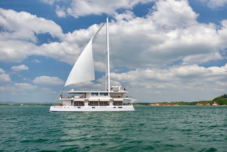 A large white catamaran yacht with a raised sail glides on calm blue-green water under a partly cloudy sky, with a distant shoreline and greenery visible in the background.