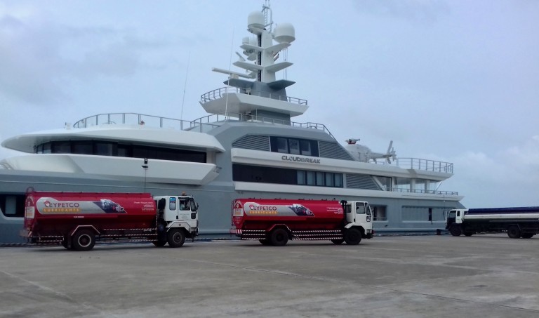 A large luxury yacht is docked at a pier while three fuel lorries, two red and one blue, are parked beside it, refuelling the vessel under an overcast sky.