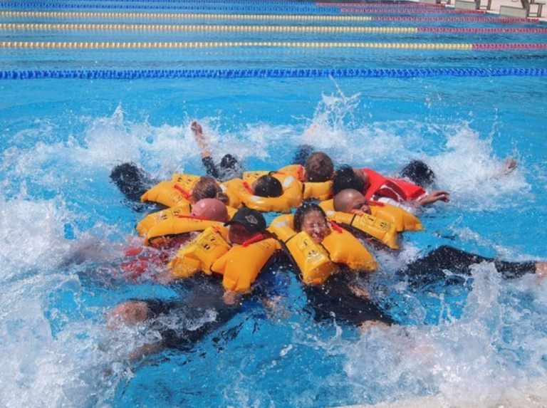 A group of people wearing bright yellow lifejackets float together in a swimming pool, forming a tight circle as water splashes around them. Colourful lane dividers are visible in the background.