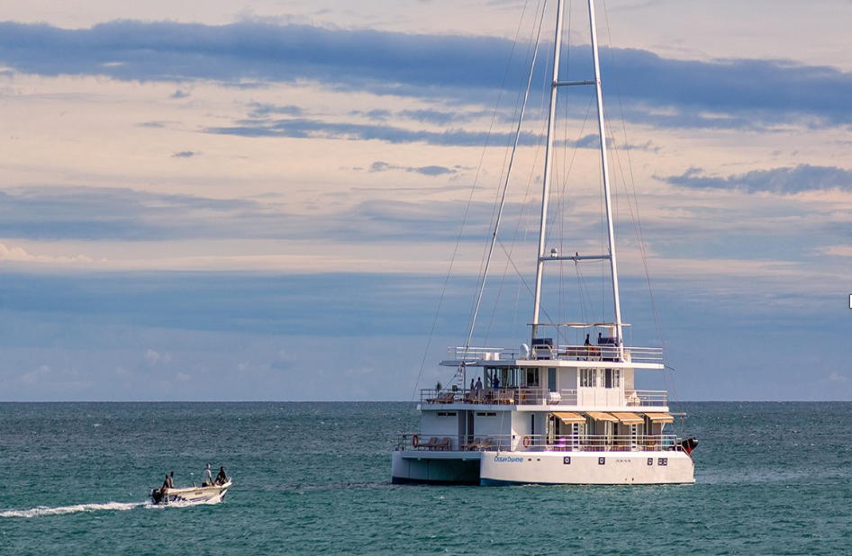 A large white catamaran is anchored on calm turquoise water under a cloudy sky, with a small motorboat carrying several people approaching from the left.