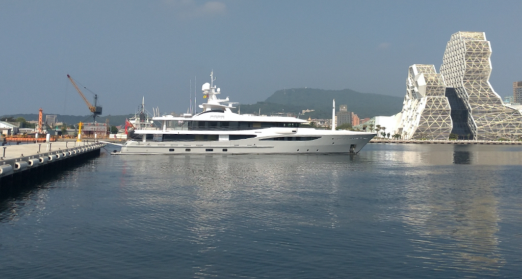 A large white luxury yacht is moored at a harbour with calm water, modern architectural buildings, and a crane in the background under a clear blue sky.