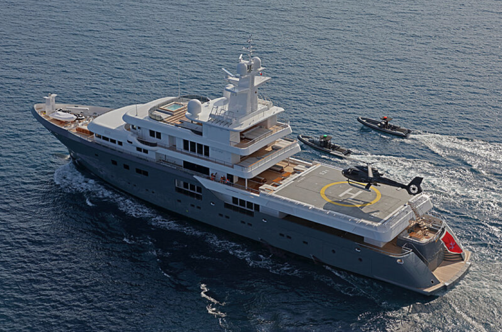 A large luxury yacht with a helipad and helicopter on deck sails on the sea, accompanied by two smaller speedboats.