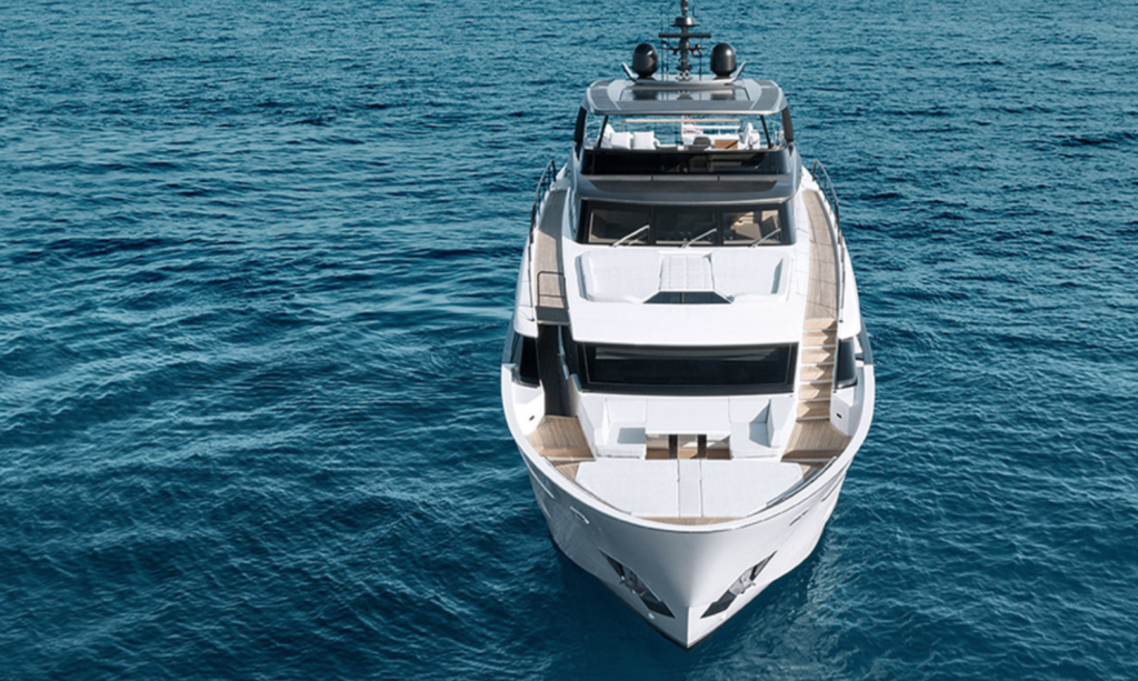 A sleek white yacht is seen from above, cruising on calm, deep blue ocean water on a clear day. The vessel features multiple decks and modern design details.