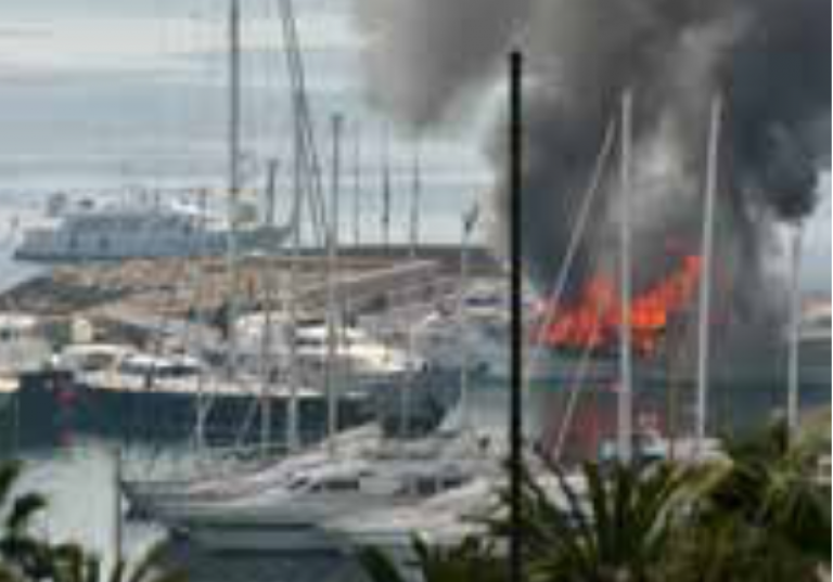 A marina with several docked yachts; one boat is on fire, producing thick black smoke and visible flames, while other boats remain unaffected nearby.