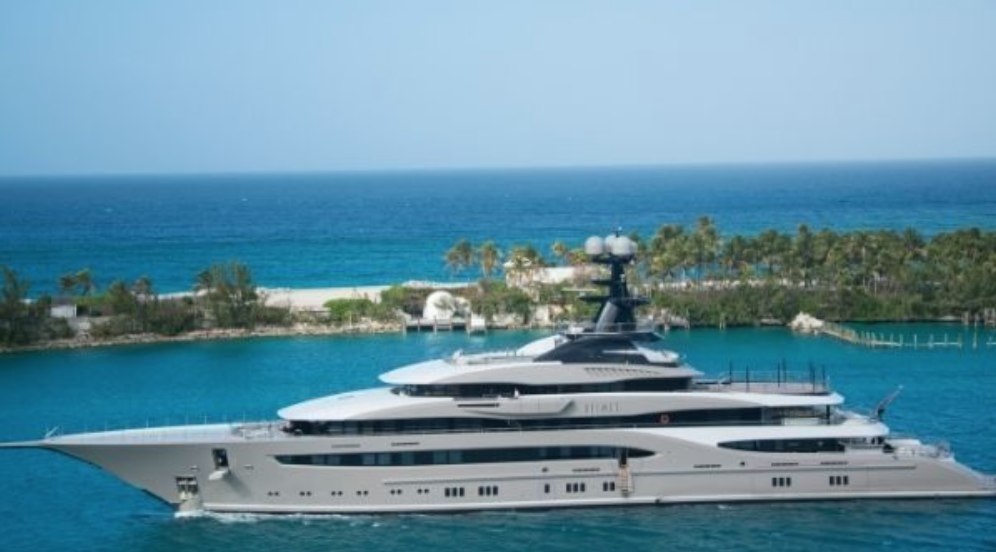 A large, modern luxury yacht sails on clear blue water near a tropical shoreline lined with palm trees under a bright, sunny sky.