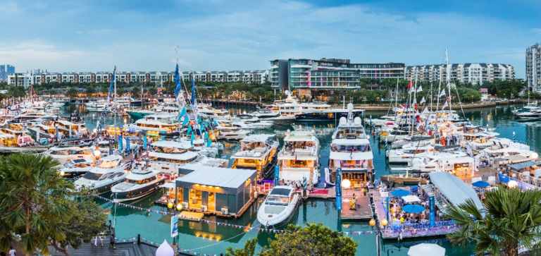 A bustling marina filled with numerous yachts and boats, surrounded by modern buildings and palm trees, under a cloudy blue sky during twilight. People are seen walking along jetties decorated with lights and flags.