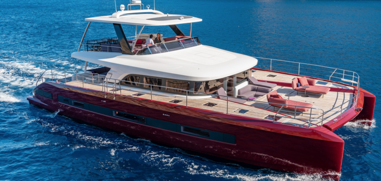 A modern red and white luxury catamaran yacht with sun loungers and seating areas cruises on clear blue water, with several people relaxing on the upper deck.