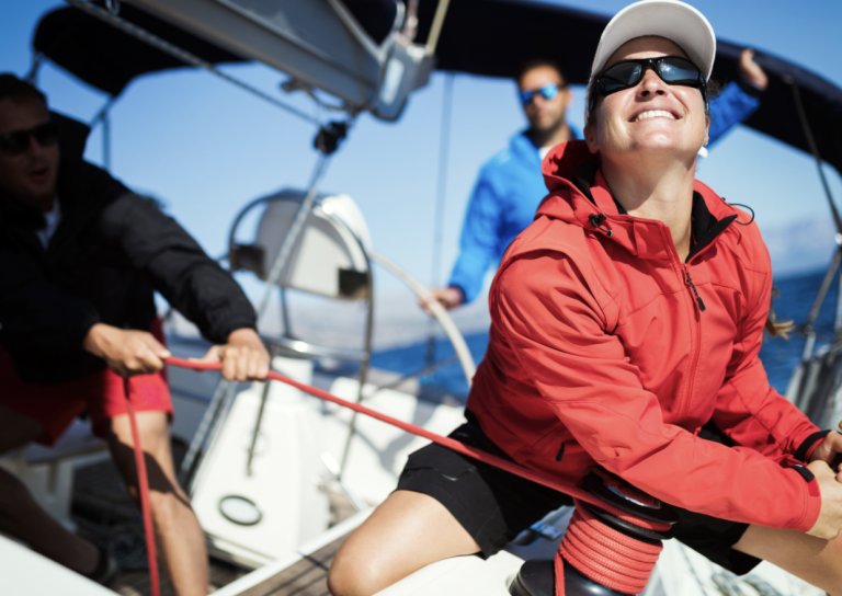 Three people on a sailboat work together, pulling ropes and smiling, with blue sky and sea in the background. The person in front wears a red jacket, cap, and sunglasses, appearing focused and joyful.