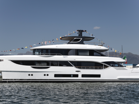 A large, modern white yacht decorated with colourful flags is moored on calm water under a clear blue sky, with distant mountains visible in the background.