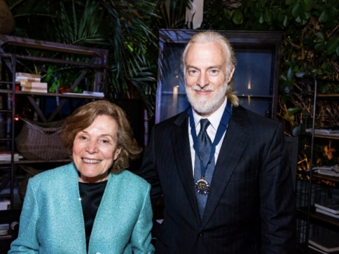 An older woman in a turquoise jacket stands smiling beside a man in a black suit and tie with a medal round his neck, both posing indoors with plants and shelves in the background.