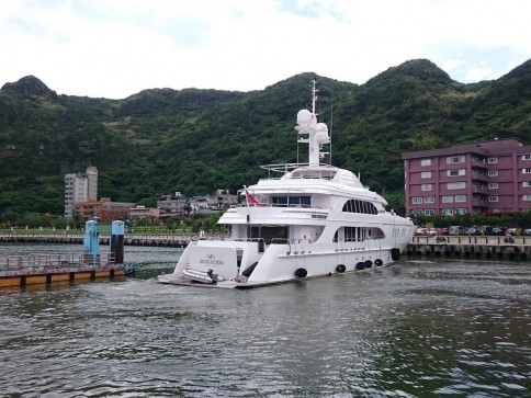 A large white yacht is docked in a harbour with green, tree-covered hills in the background and several buildings along the shore. The water is calm and the sky is partly cloudy.