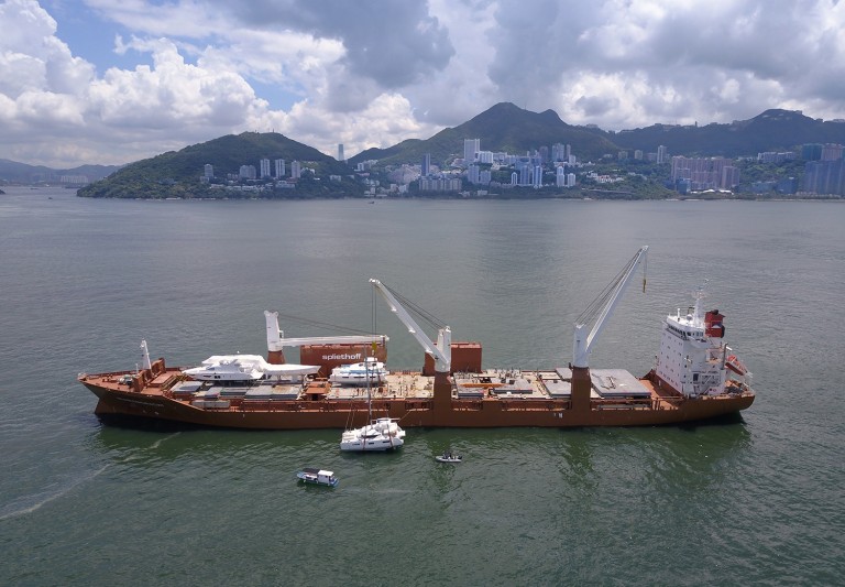 A large cargo ship with cranes is anchored in a bay near a city with high-rise buildings and green hills. Small boats are nearby on the water under a partly cloudy sky.