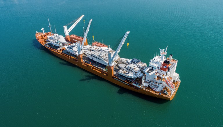 An aerial view of a large cargo ship transporting several yachts on its deck, with cranes positioned for loading and unloading, floating on calm blue water.