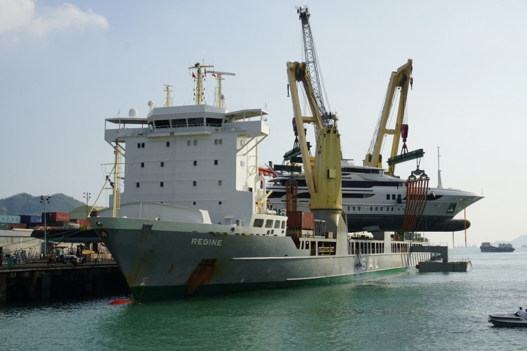 A large cargo ship named REGINE is docked at a port while cranes load or unload a luxury yacht from its deck. The scene takes place near the water with other vessels and land visible in the background.