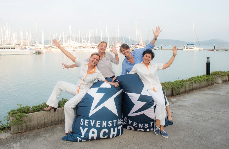Four smiling women pose with arms raised on large beanbags labelled SEVENSTAR YACHT, by a marina with many sailing boats and calm water in the background. The atmosphere is cheerful and relaxed.