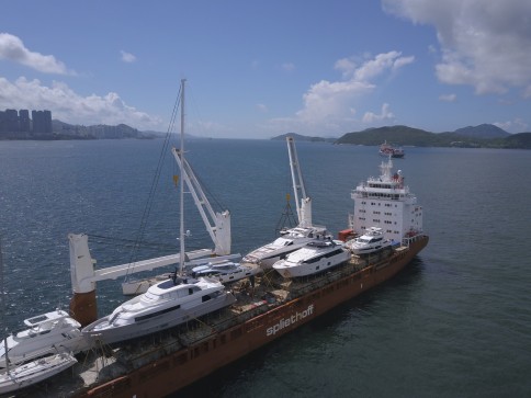 A cargo ship carrying several large yachts on its deck sails across a calm sea, with city buildings, green hills, and a partly cloudy sky in the background. The ship has cranes and the name spilitchoff visible.