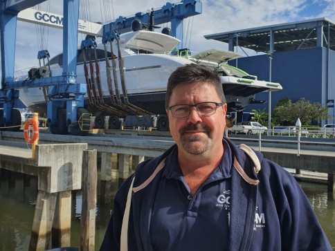 A man wearing glasses and a navy blue jacket stands on a quay in front of a large yacht being lifted by a blue crane at a marina. Industrial buildings and clear skies are visible in the background.