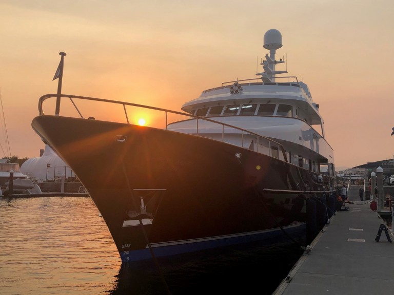 A large yacht is moored at a marina during sunset, with the sun low in the sky and casting a warm glow over the water and vessel. The yacht’s bow faces forward, and the quay is visible to the right.