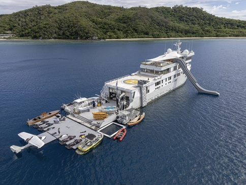 A large luxury yacht anchored near a lush green island, with a waterslide, helipad, and multiple smaller boats, jet skis, and water toys arranged on a floating pontoon beside it.
