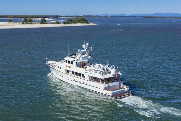 A white luxury yacht cruises through blue water near a sandy shoreline with trees, under a clear sky. Mountains can be seen in the distance.