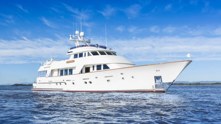A large white luxury yacht floats on calm blue water under a bright, partly cloudy sky. The yacht features multiple decks and modern amenities. Distant shoreline and hills are visible in the background.