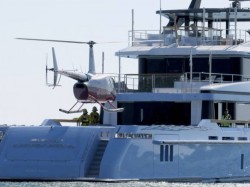 A helicopter is landing on the deck of a large, modern luxury yacht on the water. Two people in yellow vests stand nearby on the yacht’s deck.