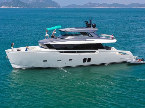 A modern white yacht floats on clear blue water with several people on board, some enjoying the sun and one waving a cloth. Hills and coastline are visible in the distance under a clear sky.