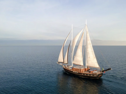 A large sailboat with three white sails glides across calm, open water under a clear sky, with no land in sight.