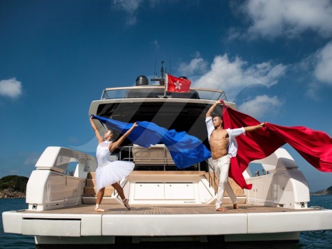 A man and woman dance on the deck of a yacht, holding flowing red and blue fabric. The sky is blue with scattered clouds, and the yacht is anchored near a coastline.