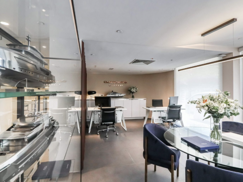 A modern office with sleek furniture, white desks, blue chairs, a round glass table with a bouquet of white flowers, and model yachts displayed on shelves. Soft lighting creates a clean, professional atmosphere.