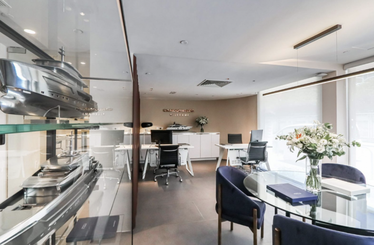 Modern office with glass walls, white desks, navy blue chairs, and a round glass table topped with a bouquet of white flowers. Shelving with metal model boats decorates the left side. Large windows let in natural light.