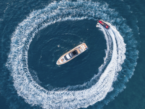 Aerial view of a yacht floating in the ocean whilst a smaller speedboat circles it, creating a large ring of white wake on the blue water.