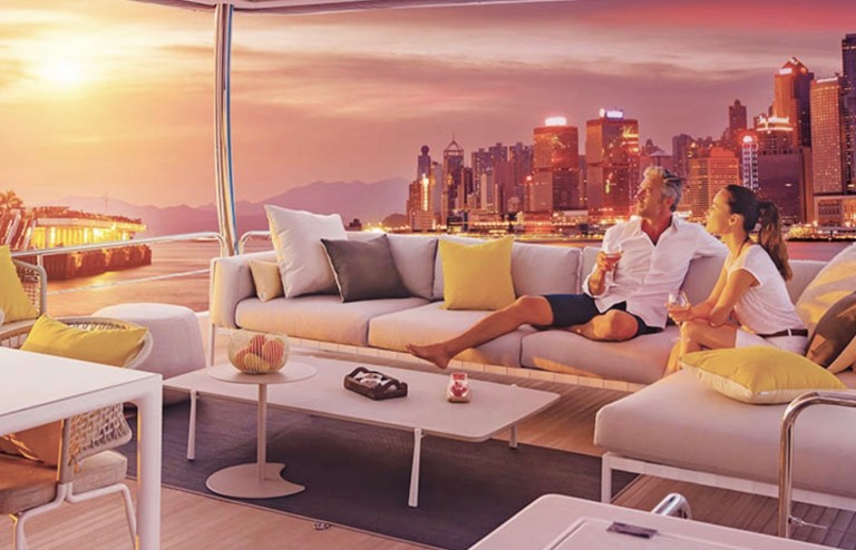 A couple relaxes on a yacht at sunset, enjoying drinks on a modern terrace with city skyscrapers and a colourful sky in the background.